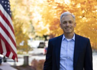 John Walsh Wins Denver DA Primary, Other Races Set for November John Walsh, the victor in Denver’s District Attorney primary, stands outside in a blue suit jacket with an American flag and trees with fall leaves behind him.