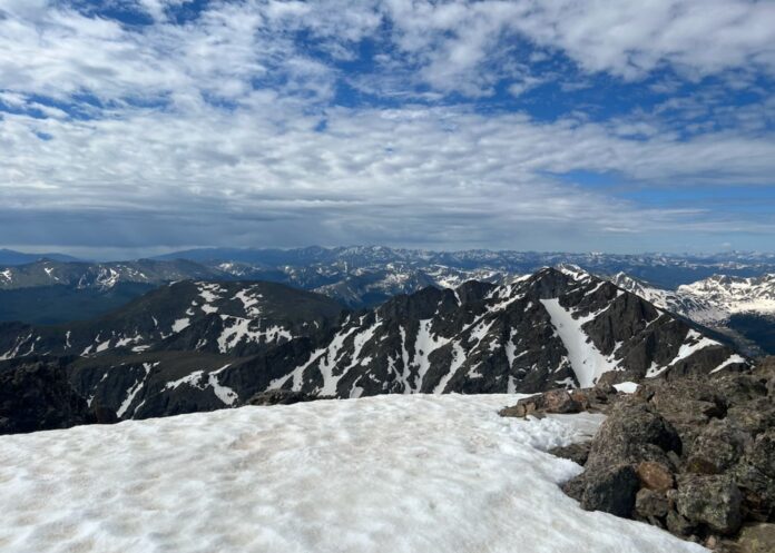 Resize image template - Editorial A photo from the summit of Mount of the Holy Cross. Snow from the peak is in the foreground, with many other mountains from the Sawatch range visible in the background under a partly cloudy sky.