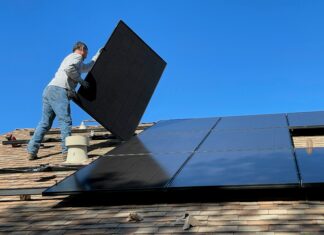 Legislative Effort to Expand Automatic Residential Solar Permitting Clears First Hurdle A man holds a solar panel on a tiled roof and works to install it next to several already installed solar panels.