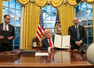 Trump Targets Law Firms with Ties to Political Opponents in Series of Executive Orders President Trump, sitting, shows a signed executive order to the camera while in the Oval office and flanked by two advisors.