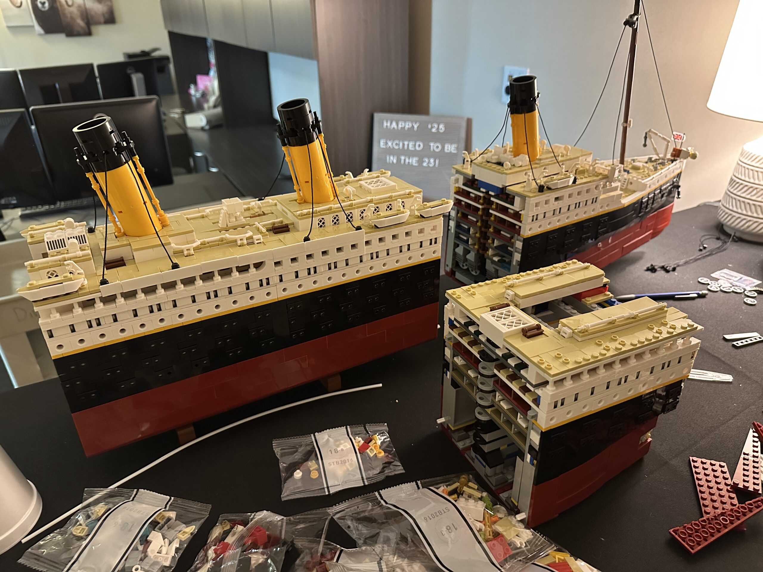 A Lego model of the Titanic rests on a desk in big pieces. It’s in the process of being assembled. Just behind the model, a sign reads “Happy ‘25 Excited to be in the 23!”