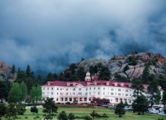 A Frozen Dead Guy, Ghosts, Hollywood Artifacts: The Sale of the Stanley Hotel The Stanley Hotel, a large white building with a red roof, sits in front of a hillside with dark clouds roiling above.